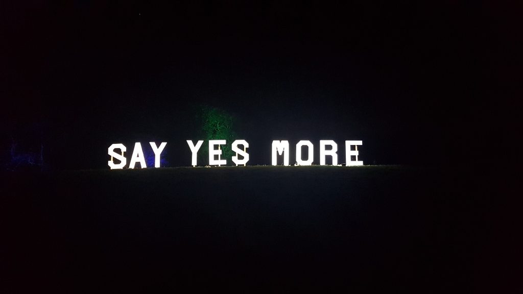 A sign saying "say yes more", white lights against the darkness
