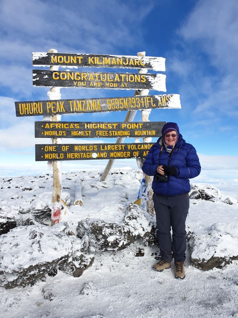 Me in front of sign at Uhuru point, summit of Kilmanjaro, heght 5895. Snow on ground and blue skies