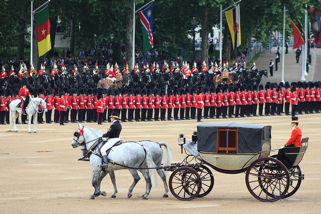 A carriage pulle dby two grey horses, making its way across Horse Guards parade in front of a line of red jacketed soldiers