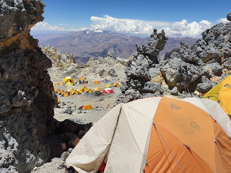 Orange and white tent set up at edge of cliff with more tents on plateau below. Mountains in the distance