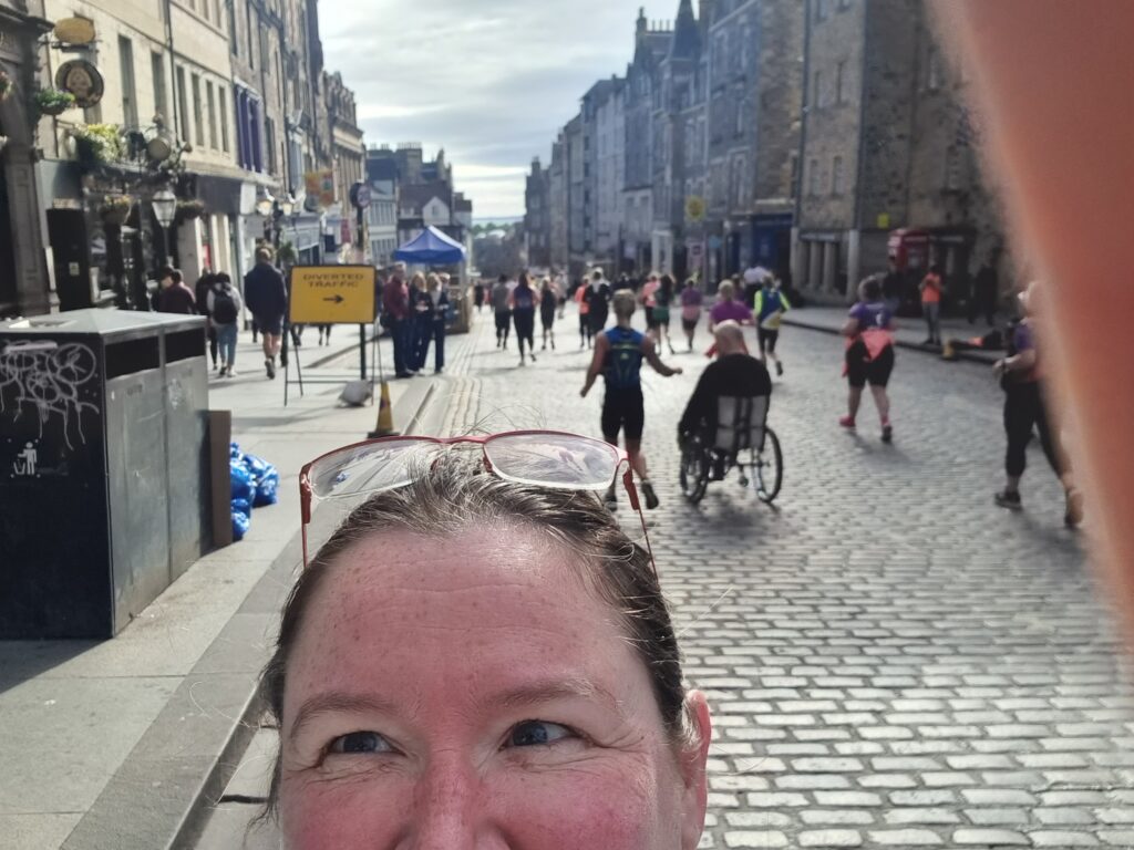 Very bad selfie with only half a face showing; me on the cobbled street of the Royal mile with runners heading down the hill