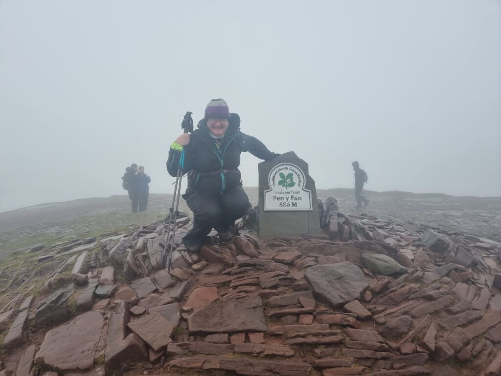 Crouching at the summit marker on Pen Y Fan, 886m high