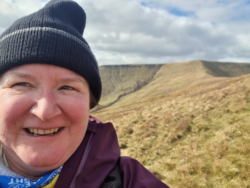 selfie with me in a wooly hat with a hill ridge behind me