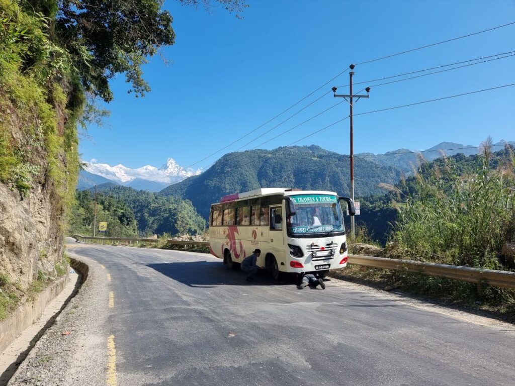 Picture of bus at side of road, man under neath trying to change tyre. Hills and mountains in the background