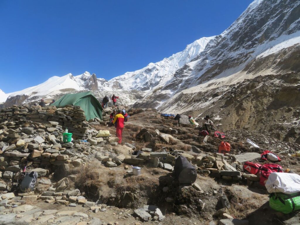 rocky campsite with porters and bags. Only kitchen tent is set up, befind are snowy slopes