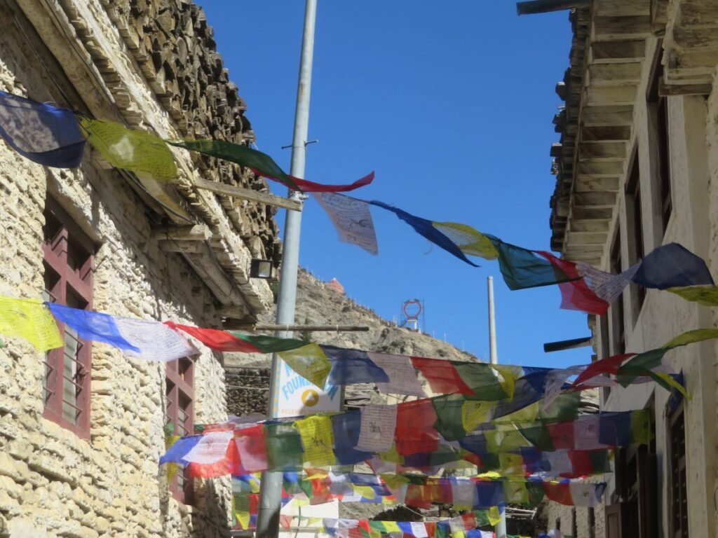 Narrow street, whitewashed buildings, prayer flags. The Marpha Apple sign on hill in background
