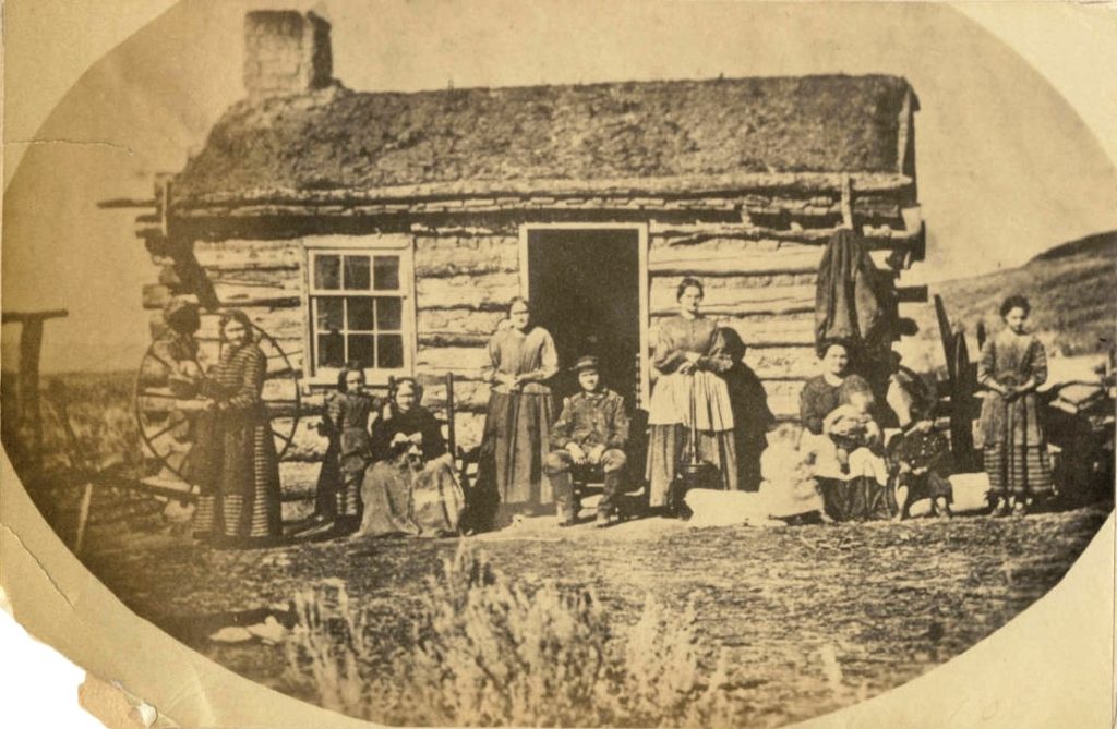 sepia photo, taken in Kaysville, Utal May 10th 1869. There are a group of people, sitting and standing in front of a log cabin.  